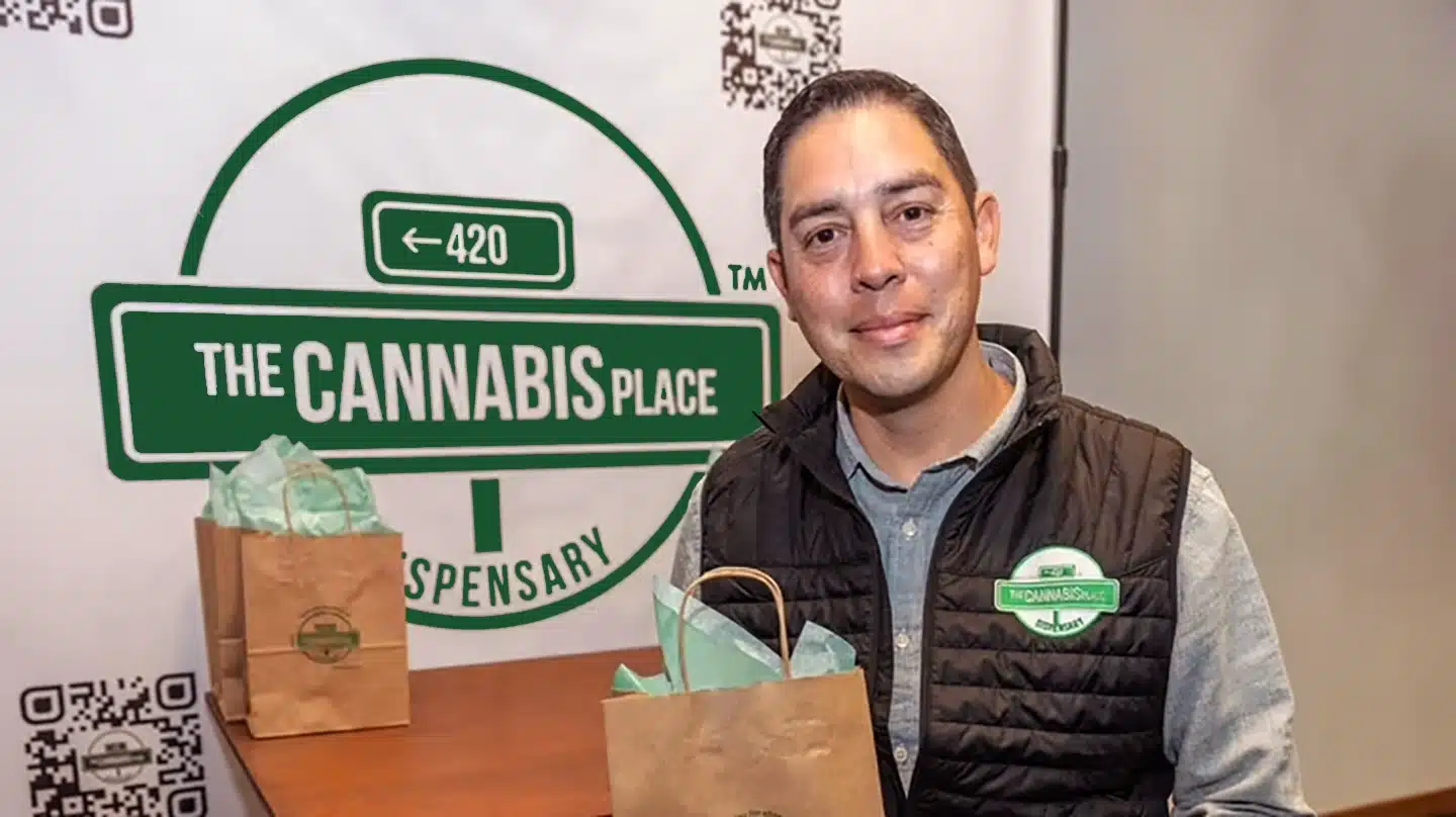 A man stands indoors holding a paper bag in front of a banner that reads The Cannabis Place Dispensary.