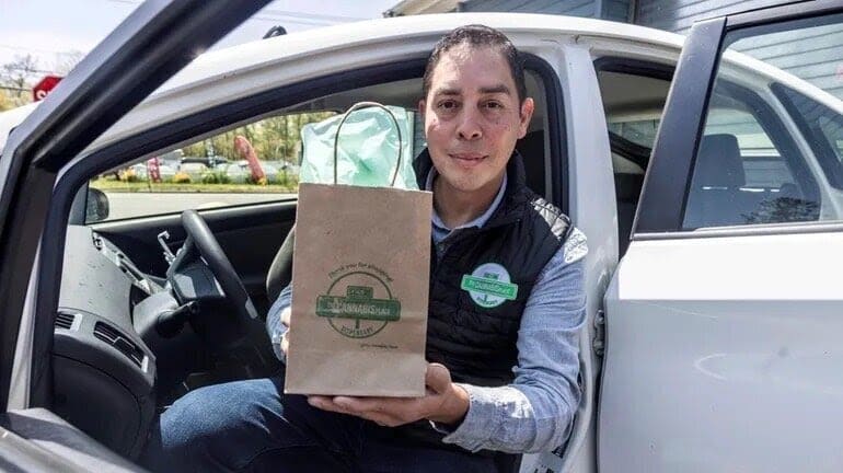 A man sits in a car with the door open, holding a brown paper bag with a logo, suggesting a takeaway or delivery service.