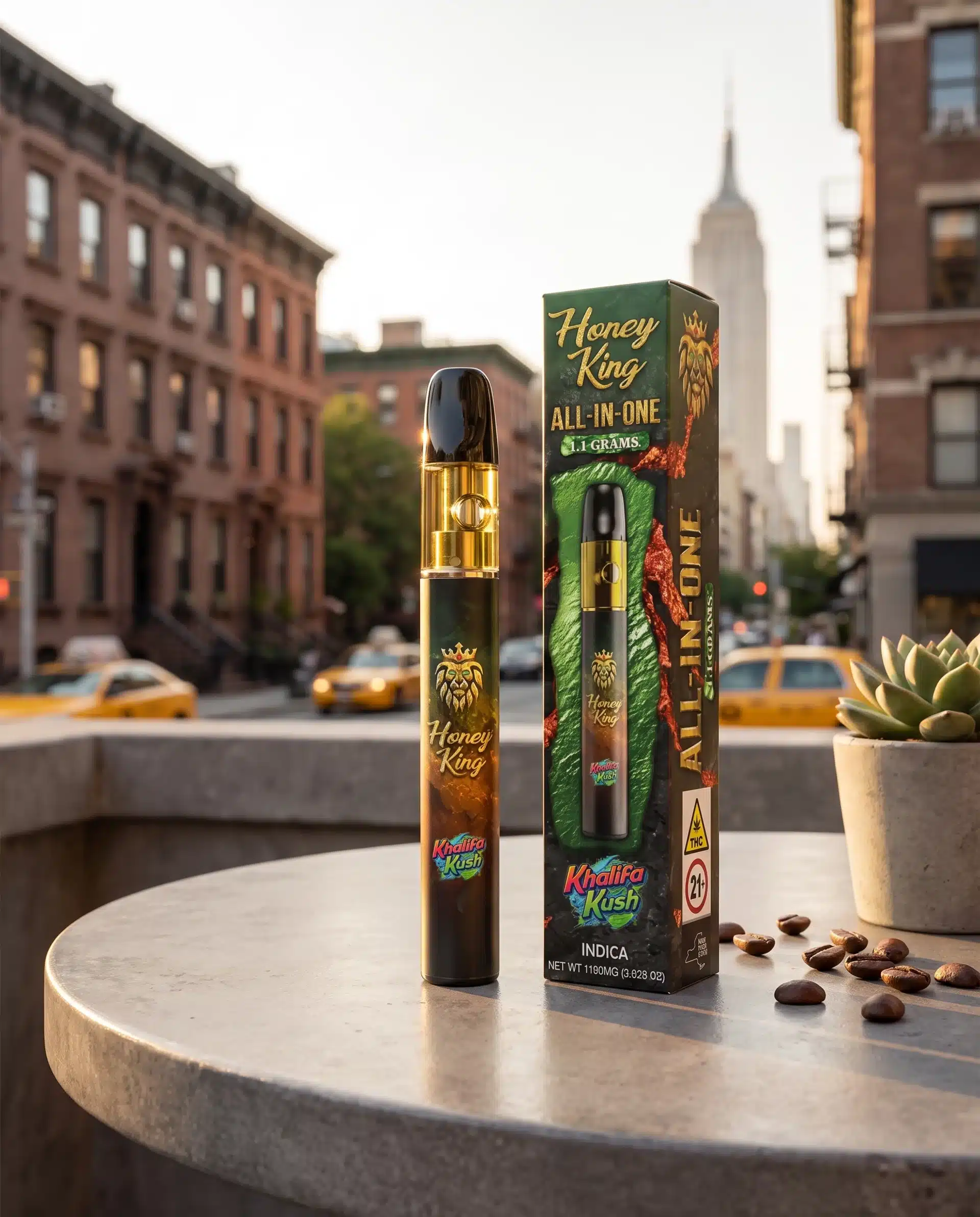 A Honey King vape pen and its box labeled Kushika Kush Indica on a round table with a succulent plant, coffee beans, and New York City buildings and taxis in the background.